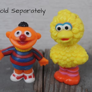 May include: Two colorful figurines of children's characters. One is orange with a red nose and black hair, wearing a striped shirt. The other is a large yellow bird with orange feet and a red beak. The text "*sold separately" is in the image.
