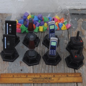 May include: A collection of retro-themed miniature toys, including black cassette tapes with "AWESOME MIX" labels, lava lamps, cell phones, and joysticks. A bag of colorful geometric shapes is in the background. The toys are on a weathered wooden surface.