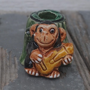 Könnte beinhalten: Keramik-Affenfigur, die eine gelbe Gitarre spielt, vor einem grünen und schwarzen Baumstumpf. Der Affe hat ein lächelndes Gesicht und trägt eine schwarze Jacke. Die Figur steht auf einer verwitterten Holzoberfläche.