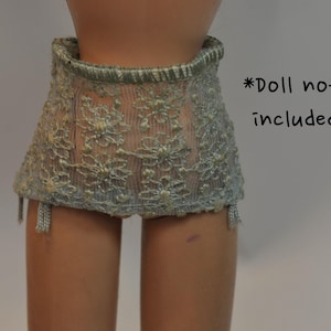 May include: A light blue doll skirt with floral lace detailing. The skirt has a scalloped edge and attached garter straps. The text "*Doll not included" is written in black.