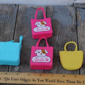 May include: A selection of Barbie accessories: three pink tote bags with a puppy design and the Barbie logo, a turquoise square bag, and a yellow woven basket. A wooden ruler is at the bottom.