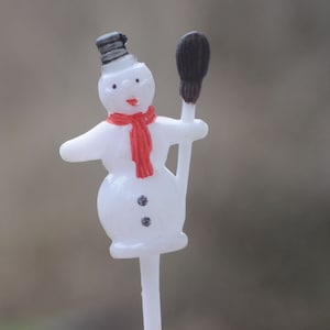 May include: A small, white snowman figurine with a black top hat, red scarf, and black coal buttons. The snowman holds a black broom and is mounted on a white stick, ideal for festive decorations.