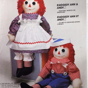 May include: Two Raggedy Ann and Andy dolls. One doll wears a white apron with red trim over a floral dress. The other doll wears a red and white checkered shirt, blue pants, and a sailor hat. The text reads "Raggedy Ann & Andy... Traditional Favorites for Generations."