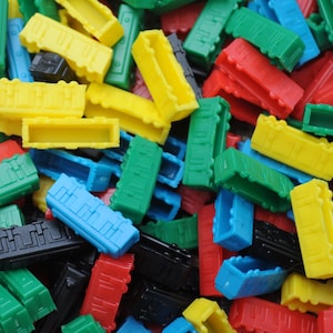 May include: A collection of small, rectangular plastic game pieces in various colors including yellow, green, red, blue, and black. Each piece features a hollow center and molded structural details on the exterior sides, resembling miniature train cars.