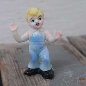 May include: A small porcelain figurine of a young child with blonde hair, wearing light blue overalls and black shoes. The figurine has its arms raised and a surprised expression. The figure is standing on a weathered wooden surface.