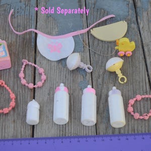May include: A collection of pink and white baby doll accessories, including a bib, bottles, rattles, and necklaces. *Sold Separately*