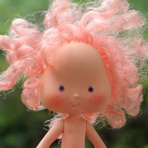 May include: A pink plastic doll with curly pink hair, blue eyes, and a smiling face. The doll is wearing a pink dress and has a pink bow in her hair.