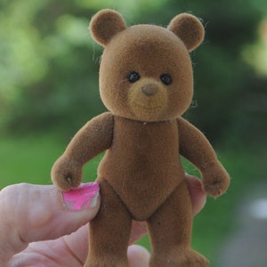 May include: A brown teddy bear with a soft, plush texture. The bear has black eyes and a black nose. The bear is standing on a hand.