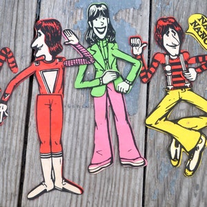 Może przedstawiać: Trzy zabytkowe papierowe wycinanki postaci z kreskówek. Jedna postać jest w czerwonym kombinezonie z białym trójkątem. Druga ma zieloną kurtkę i różowe spodnie. Trzecia postać ma czerwono-czarną koszulę w paski i żółte spodnie, z dymkiem z napisem "Na-No!"