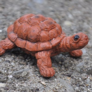 May include: A small, reddish-brown turtle figurine with a textured, segmented shell and a detailed head featuring a single visible black eye. The turtle is depicted in a walking position with four legs extended outwards, showcasing a realistic, moulded surface texture throughout its entire body.