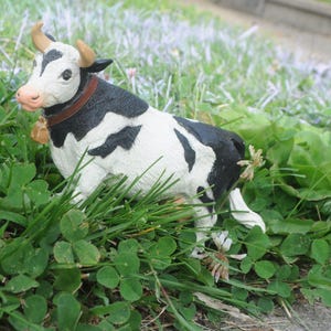 May include: A black and white cow figurine with brown horns and a bell around its neck. The cow is set in a bed of green grass and clover. The cow has a pink nose and is looking to the left.