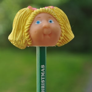 May include: A yellow plastic doll's head with blue eyes and pink bows in her hair is attached to a green pencil with the word "CHRISTMAS" printed on it.