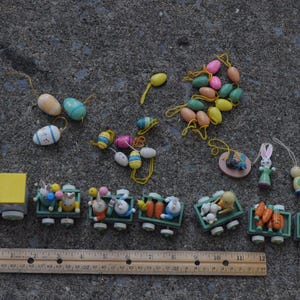 May include: A collection of miniature wooden Easter ornaments including small painted eggs, a rabbit, a bird, and a toy train with six cars carrying figures and vegetables, all arranged above a 12-inch wooden ruler for scale reference.