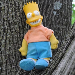 May include: A Bart Simpson doll featuring a yellow plastic head with a wide grin and spiky hair, wearing a peach-colored short-sleeved fabric shirt, bright blue fabric shorts, and matching blue fabric shoes with white trim accents.