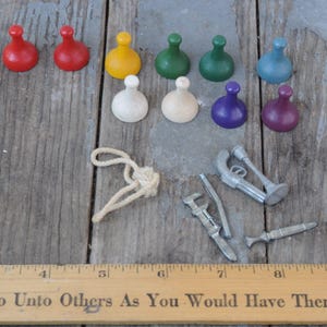 May include: A collection of vintage game pieces and tools, including red, yellow, green, blue, purple, and white game tokens. Also, a rope, a wrench, a small metal gun, and a metal knife are visible. A wooden ruler is at the bottom.