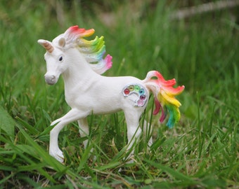 May include: A white unicorn figurine with a multi-colored rainbow mane and tail. The unicorn features a silver-colored artist palette symbol on its flank, decorated with small, colorful circular gems. It has a single horn on its forehead.