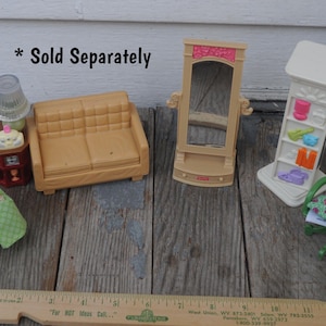 May include: A selection of miniature dollhouse furniture, including a tan sofa, a green chair, a mirror, a white shelf unit, and a nightstand with a lamp. The items are arranged on a weathered wooden surface.