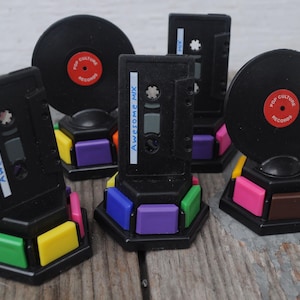 May include: A selection of black cassette tape and vinyl record holders with colourful bases. The vinyl records have red labels that read "Pop Culture Records." The cassette tapes have the text "Awesome Mix" on them.