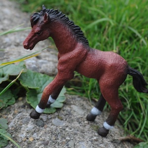 May include: A brown and black toy foal stands on a gray stone surface, surrounded by green grass and leaves. The foal has a dark mane and tail, with white markings on its legs. The toy is a detailed representation of a young horse.