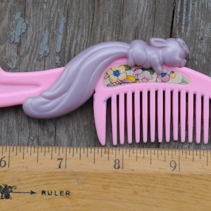 以下が含まれることがあります： 装飾的なデザインのピンク色のプラスチック製の櫛。櫛は、薄紫色の尾と小さなウサギのフィギュアが特徴です。櫛のハンドルはユニークな形をしています。櫛の長さは約10cmです。