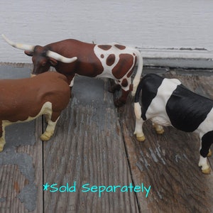 May include: Three toy farm animals: a brown bull with white horns, a brown and white spotted longhorn, and a black and white cow. The toys are on a weathered wooden surface.