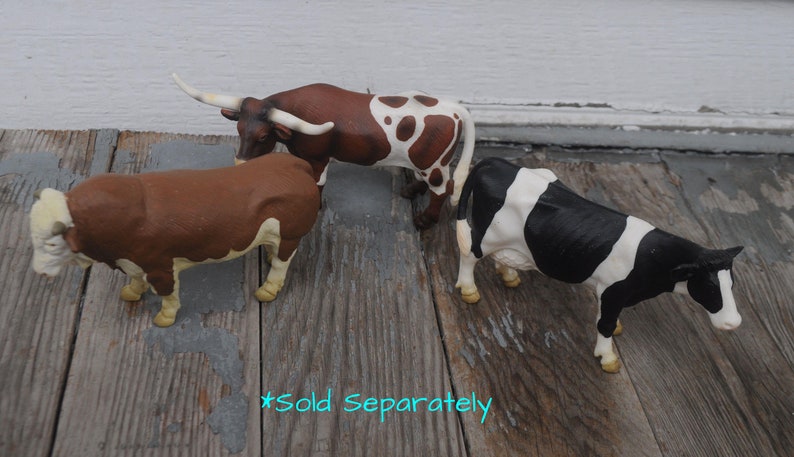 May include: Three toy farm animals: a brown bull with white horns, a brown and white spotted longhorn, and a black and white cow. The toys are on a weathered wooden surface.