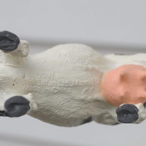 May include: A small, white and black cow figurine with a pink udder and black hooves. The underside of the toy is visible, with text that reads "COW" and "Made in China". The cow is being held by a person.
