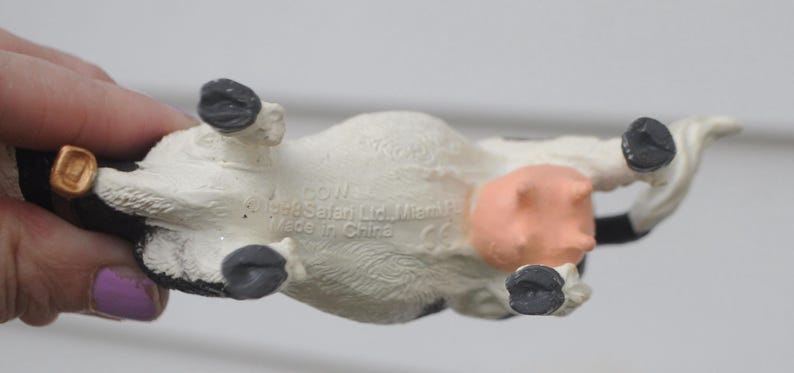 May include: A small, white and black cow figurine with a pink udder and black hooves. The underside of the toy is visible, with text that reads "COW" and "Made in China". The cow is being held by a person.