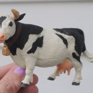 May include: A small, decorative cow figurine with a black and white pattern. The cow has brown horns, a bell around its neck, and a pink nose. The figurine is held in a hand.