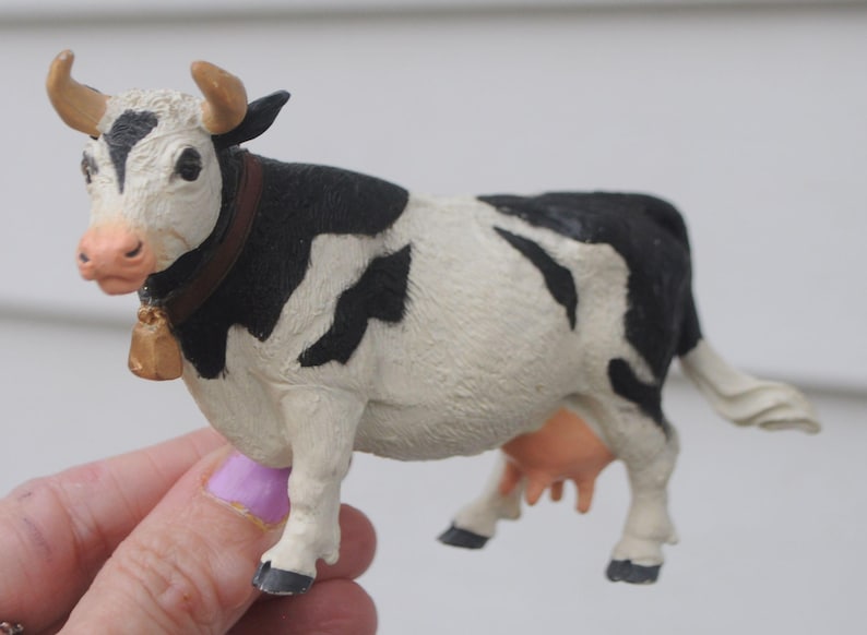May include: A small, decorative cow figurine with a black and white pattern. The cow has brown horns, a bell around its neck, and a pink nose. The figurine is held in a hand.