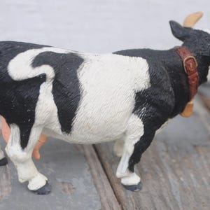 May include: A black and white cow figurine with a brown collar and bell. The cow has tan horns and is standing on a wooden surface. The figurine is a farm animal collectible.