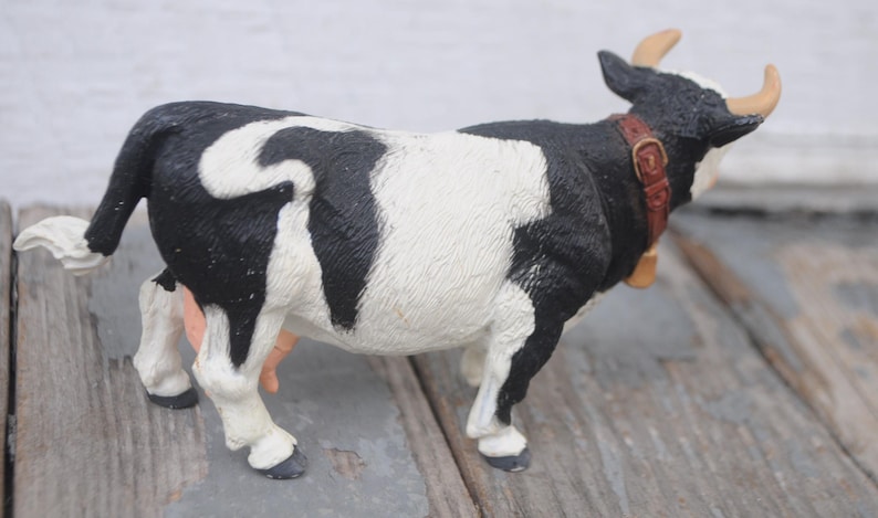 May include: A black and white cow figurine with a brown collar and bell. The cow has tan horns and is standing on a wooden surface. The figurine is a farm animal collectible.