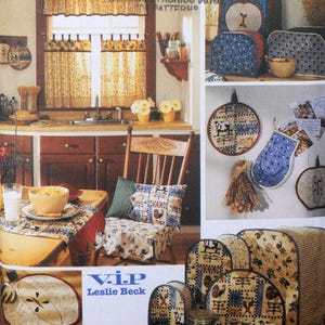 Puede incluir: Una escena de cocina de estilo rústico con varios artículos de tela. Incluye una silla con un cojín estampado, una mesa puesta y varias fundas para tostadoras. La paleta de colores incluye rojos, azules y amarillos. El texto "V.I.P Leslie Beck" es visible.