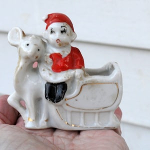 May include: A vintage ceramic Christmas planter depicting Santa Claus in a sleigh pulled by a reindeer. The planter is white with gold trim and red and black details. A charming festive decoration.