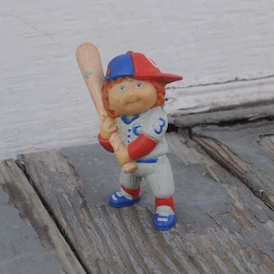 Könnte beinhalten: Eine kleine Plastikfigur eines Baseballspielers, der eine blaue und rote Baseballkappe, ein weißes und blaues Trikot mit der Nummer 3 und blaue und weiße Schuhe trägt. Die Figur hält einen hölzernen Baseballschläger.