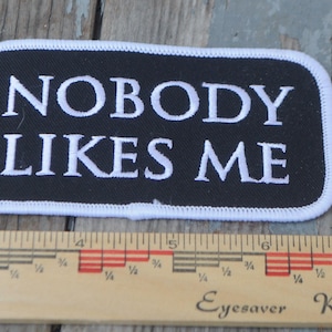 May include: Black rectangular patch with white embroidered text that reads "NOBODY LIKES ME." The patch has a white border and is placed on a wooden surface. A ruler is below the patch.
