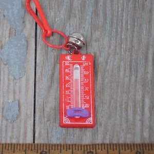 Könnte beinhalten: Ein roter Plastik-Thermometer-Schlüsselanhänger mit einem silbernen Glöckchen-Anhänger. Das Thermometer hat einen violetten Plastikring um die Glühbirne und ein weißes Zifferblatt mit schwarzen Markierungen. Das Thermometer zeigt von 10 bis 50 Grad Celsius und von 20 bis 120 Grad Fahrenheit an.