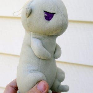 May include: A light gray plush toy in the shape of a small creature, with a purple eye and a white string attached. The toy is held in a person's hand. The plush has a stitched design.