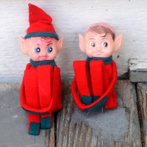 May include: Two vintage elf dolls. Each doll has a plastic face with painted features, including blue eyes. They wear red felt outfits with green trim, including hats and collars. The dolls are posed on a weathered wooden surface.