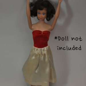 May include: A vintage doll dress featuring a red velvet bodice and a cream-colored satin skirt. The dress has a strapless design and a gathered waist. The text "*Doll not included" is visible.