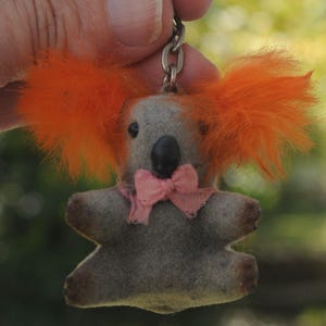 May include: A vintage koala bear keychain with orange fur accents and a pink bow tie. The koala has a gray body, black eyes, and a black nose. The keychain is attached to a silver chain.