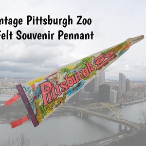 May include: Vintage Pittsburgh Zoo felt souvenir pennant. The pennant is white with colourful illustrations of zoo animals and the word "Pittsburgh" in red. The pennant has a red and purple border and red streamers.
