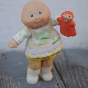 May include: A vintage Cabbage Patch Kids doll with a bald head, green eyes, and rosy cheeks. The doll wears a white dress with yellow floral accents and a green bow. It holds a small orange puppet. The doll is a collectible toy.
