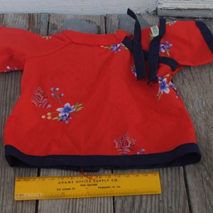 May include: Red short-sleeved shirt with a floral print and a navy blue trim. The shirt has a tie at the neck and the text "ADAMS OFFICE SUPPLY CO. DIAL 386-0490 210 ADAMS ST. FAIRMONT, W. VA." on a yellow ruler.