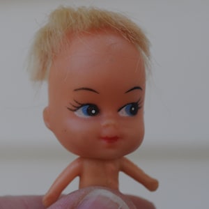 May include: A small, vintage doll with a peach-colored face, blue eyes, and blonde hair. The doll has painted facial features, including eyelashes and a red mouth. The doll is held by a person's fingers.