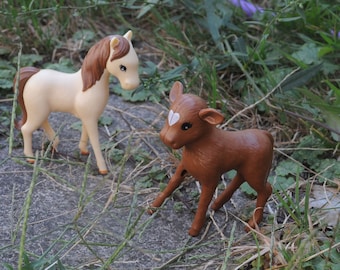 May include: A brown and white toy calf and a tan toy horse standing in a grassy area. The calf has a white heart on its forehead.