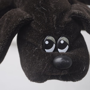 May include: A close-up of a brown plush dog toy with large, expressive eyes and a black nose. The toy has floppy ears and is made of a soft, textured material. The dog's eyes are large and shiny, with white highlights.