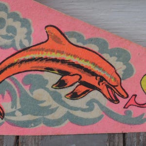 May include: A vintage pennant with a pink background. It features an orange dolphin leaping over blue waves, with the word "SEA" in a retro font. The pennant has a red border.