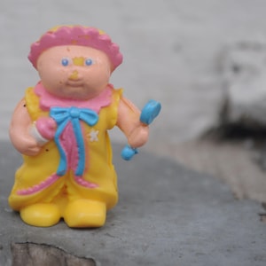 Puede incluir: Una figura vintage de Cabbage Patch Kids. La muñeca lleva una bata amarilla con ribetes rosas y un lazo azul. Tiene un gorro rosa y sostiene un biberón y una sonaja azul. La figura es de plástico.
