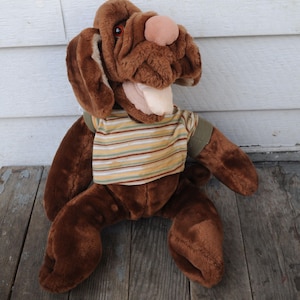 May include: A brown plush dog toy with a light pink nose and red eyes. The dog is wearing a striped shirt with yellow, brown, and green stripes. The toy is sitting on a wooden surface.